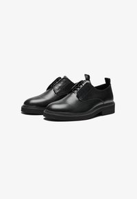 Zapatos de cuero negro con una textura suave, punta redonda y cordones elásticos para un fácil calce. Suela plana con un sutil detalle de dibujo.