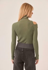 Suéter de cuello alto verde con canalé y cortes en los hombros, combinado con jeans oscuros de tiro alto en gris que cuentan con una cinturilla texturizada.