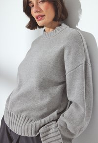 Vrouw met kort bruin haar, gekleed in een lichtgrijze breisel trui, leunend tegen een witte muur in zacht natuurlijk licht.