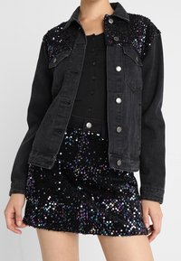 Musta denimpöytä takki, jossa on timantti koristeltu olkapäät, yhdistettynä mustaan nappipaitaan ja mustaan helmikirjailtuun hameeseen, jossa on eläviä korostuksia.