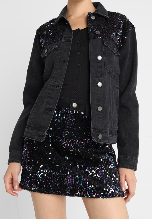 Musta denimpöytä takki, jossa on timantti koristeltu olkapäät, yhdistettynä mustaan nappipaitaan ja mustaan helmikirjailtuun hameeseen, jossa on eläviä korostuksia.