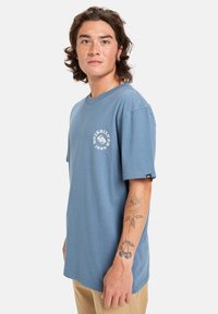 Mladý člověk s vlasy střední délky, oblečený v modrém tričku Quiksilver a béžových kalhotách, ukazuje tetování na předloktí s třešněmi a listy.