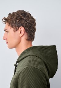 Sudadera con capucha en verde oliva, con un patrón texturizado de rombos, capucha ajustable con cordón y una silueta ceñida.