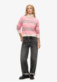 Pull en tricot avec un large col roulé, dans des motifs géométriques rose, blanc et gris, associé à un jean gris ample et des chaussures noires.