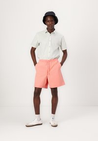Kortærmet, lysegrøn stribet skjorte med knaplukning, parret med koralrøde shorts og hvide sneakers. En sort bøttehat bæres.