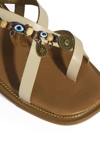 Tan leren sandaal met beige banden, decoratieve kralen en gouden muntaccenten. Platte zool met een ronde neus. Opvallende oogvormige kralen.