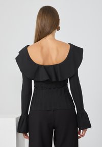 Svart strikket topp med avtagbar ruffle på skuldrene, lange ermer og plisserte detaljer; sammen med høyhoydede svarte bukser.