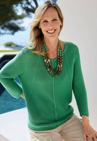 Groene gebreide trui met driekwartmouwen, voorzien van een V-hals en een textuurpatroon, gecombineerd met een multi-gekleurde kralenketting.