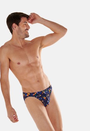 Lächelnder oberkörperfreier Mann posiert in bunten gemusterten Badehosen vor einfachem hellem Hintergrund.