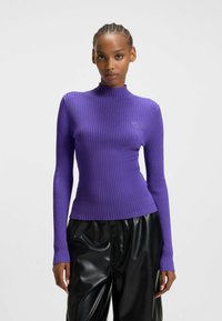 Lila gerippter Pullover mit hochgeschlossenem Kragen und langen Ärmeln, mit figurbetontem Schnitt und dezentem Logo-Detail auf der Brust, getragen mit schwarzen Hosen.