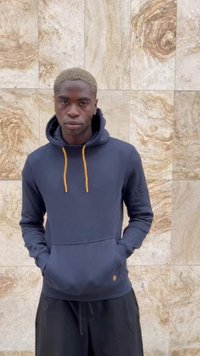 Marineblaue Kapuzen sweatshirt mit orangefarbenen Kordeln, Fronttasche und einem kleinen Logo-Detail am Saum, getragen mit schwarzen Hosen.
