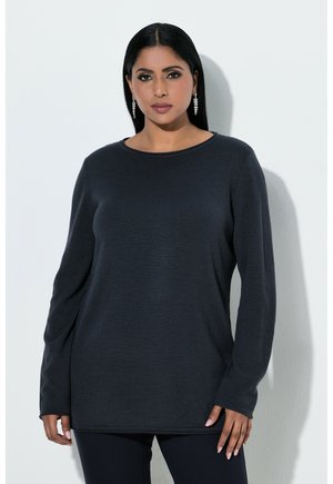 Kvinde med glat sort hår, iført langærmet mørkegrå sweater og dinglende øreringe, stående foran en ensfarvet lys baggrund.