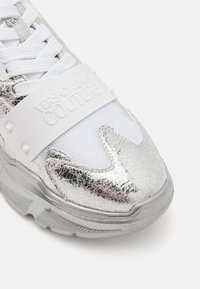 Sneakers argento con finitura metallica screpolata, dettagli in rete bianca, suole spesse e una fascia bianca con il logo "VERSACE JEANS COUTURE".