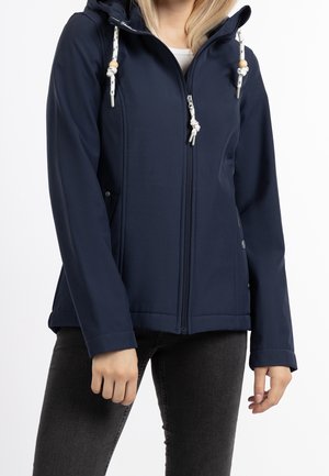 Mujer con chaqueta azul marino con capucha y cordones blancos y vaqueros negros, de pie frente a un fondo blanco.