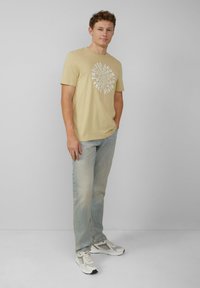 Beige katoenen t-shirt met witte cirkelvormige grafische tekst, gecombineerd met lichtblauwe denim jeans en witte sportschoenen.