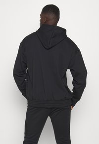 Zwarte hoodiejack met elastische zoom en manchetten, met een gladde textuur en een ontspannen pasvorm. Van achter gezien, gedragen met bijpassende broek.