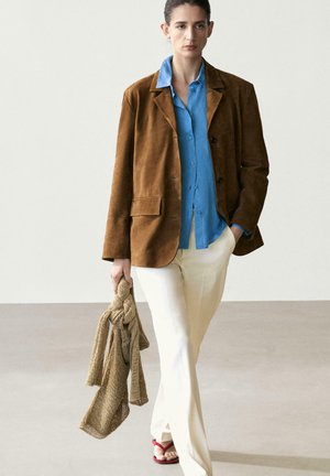 Person trägt einen braunen Blazer, blaues Hemd, weiße Hose, hält einen beigen gestrickten Schal und geht auf einem hellen Boden mit schlichtem, hellem Hintergrund.