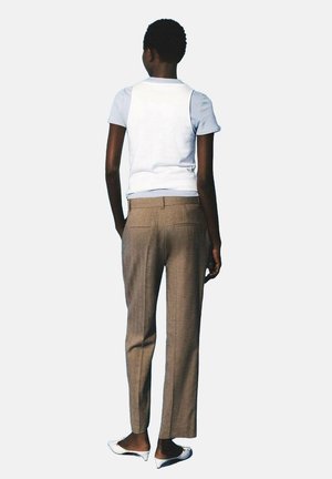 Vrouw staat met haar rug naar de kijker en draagt een witte mouwloze top over een lichtblauw T-shirt, bruine op maat gemaakte broeken en witte schoenen met een lage hak.