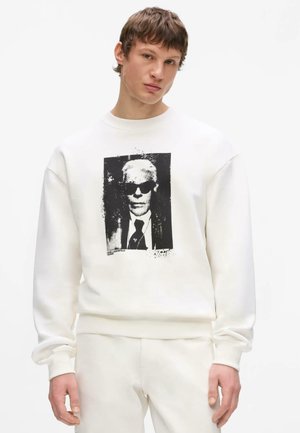 Junger Mann trägt ein komplett weißes Outfit mit einem schwarz-weißen grafischen Druck einer Person mit Sonnenbrille auf der Vorderseite des Sweatshirts.