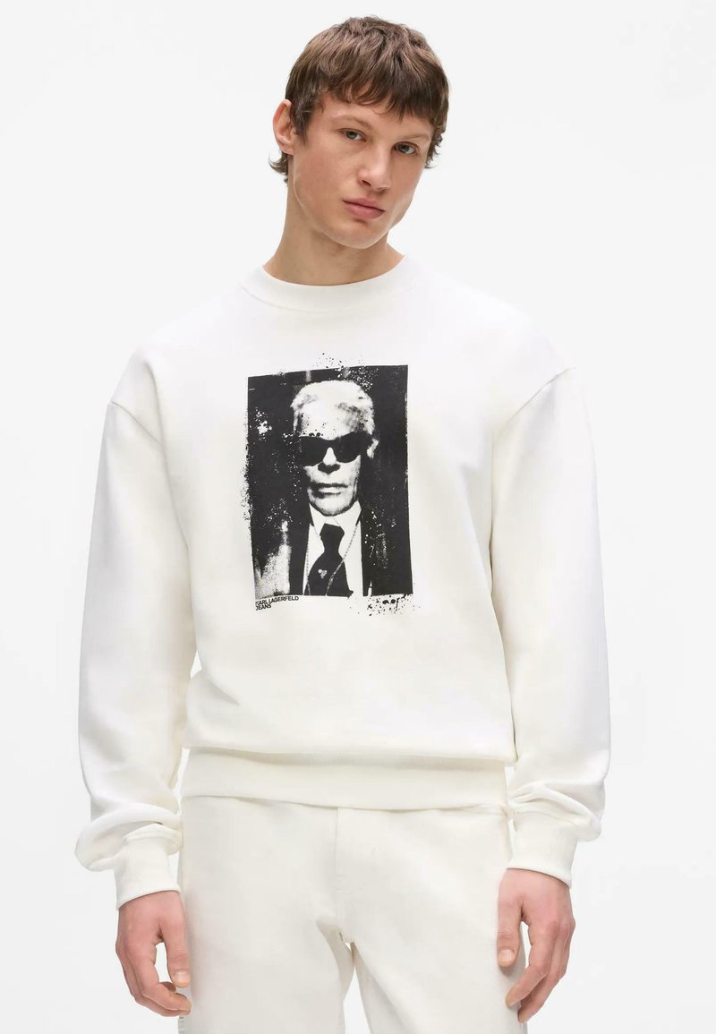 Jeune homme portant une tenue entièrement blanche avec un imprimé graphique noir et blanc d’une personne portant des lunettes de soleil sur le devant du sweat-shirt.