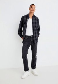 Camisa xadrez preta e azul-marinho sobre uma t-shirt branca, combinada com jeans escuros e sapatilhas brancas. Modelo está com as mãos nos bolsos.