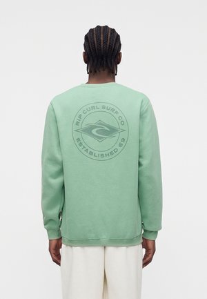 Fonott hajú személy világoszöld pulóvert visel, amelyen a hátán a "Rip Curl Surf Co Established 69" logó látható, fehér nadrággal párosítva.