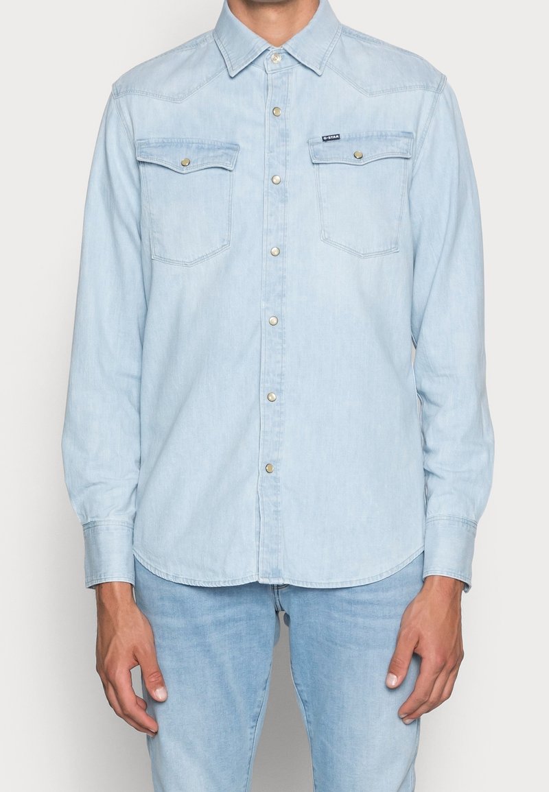 Lichtblauwe denim overhemd met lange mouwen, puntige kraag, twee borstzakken en zilveren knopen. Gladde textuur en iets gebogen zoom.