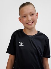 Jeune personne souriante, cheveux attachés, portant un maillot de sport noir hummel sur un fond clair uni.