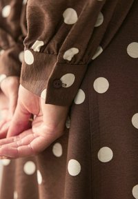 Barna pöttyös ruha, nagy fehér pöttyökkel, ráncolt ujjakkal és gombos mandzsettákkal. Az anyag textúrált, sima felülettel rendelkezik.