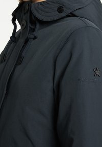 Mörkblå jacka med quiltad krage, försedd med knappar och dragsko. Diskret logotyp präglad på ärmen. Slät, lättviktig tyg.