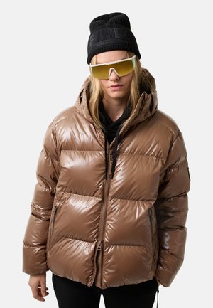 Jongere in een bruine pufferjack, zwarte beanie met de tekst "FIRE+ICE", oversized gele zonnebril, en zwarte broek voor een witte achtergrond.