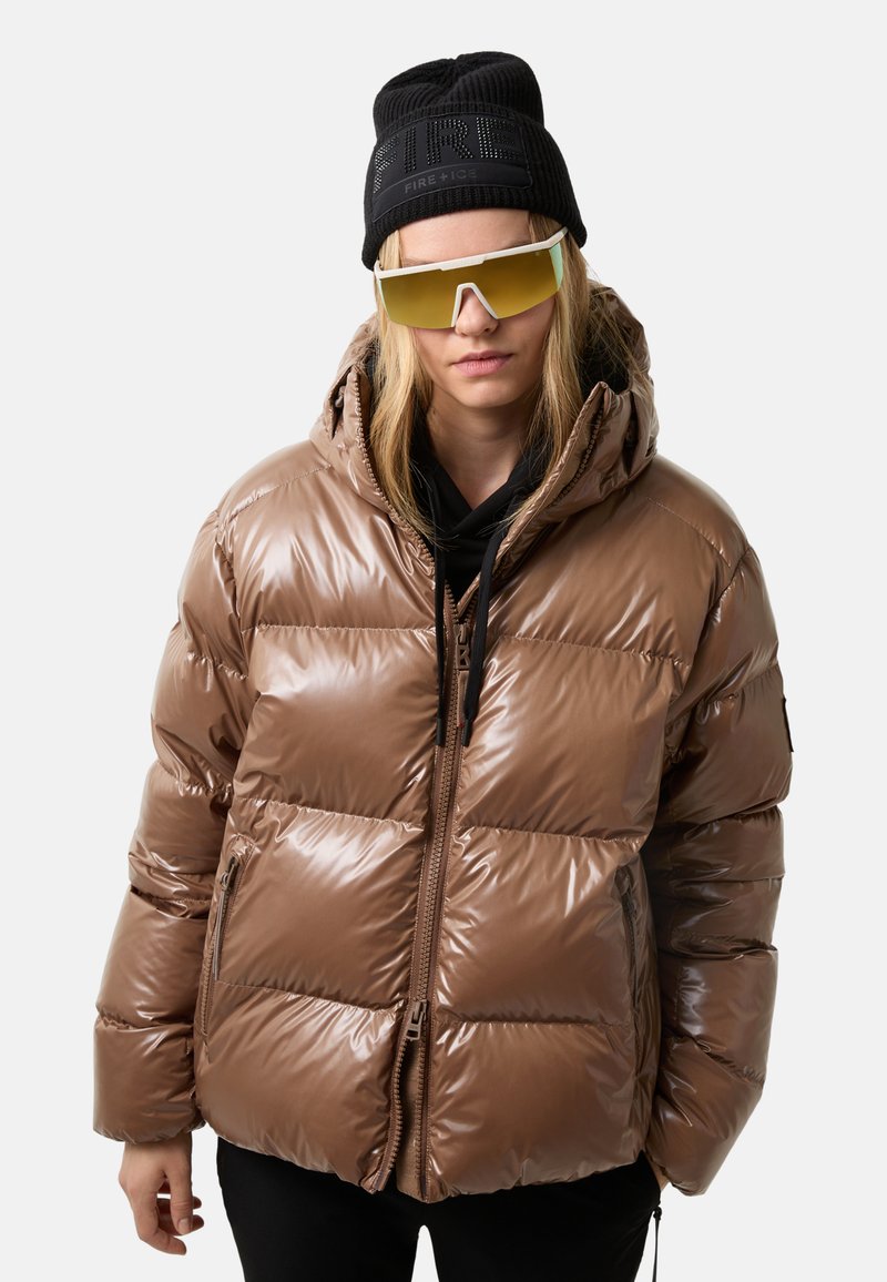 Ung person iført brun puffjakke, sort beanie med teksten "FIRE+ICE", store solbriller med gul farvetone og sorte bukser mod hvid baggrund.