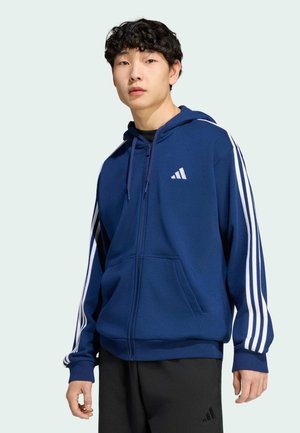Młody mężczyzna w granatowej bluzie na zamek z Adidas z białymi paskami na rękawach i czarnych spodniach Adidas, stojący na jasnym tle.