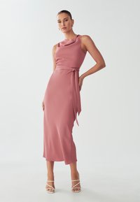 Fitted midi dress in blush pink with a cowl neckline and a side tie detail. Smooth fabric, sleeveless design, paired with strappy white heels.