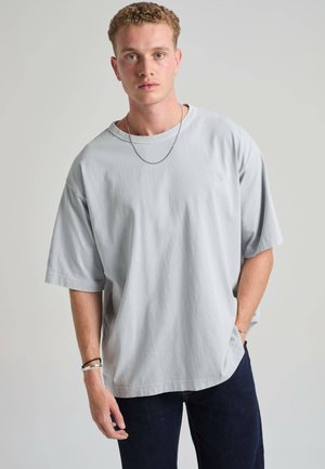 Junger Mann mit kurzen lockigen Haaren trägt ein blassgraues, übergroßes T-Shirt und dunkle Jeans, steht vor einem einfarbigen hellgrauen Hintergrund.