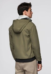 Mężczyzna w oliwkowej kurtce z kapturem z czarnymi mankietami oraz beżowych spodniach, stojący bokiem na jednolitym szarym tle.