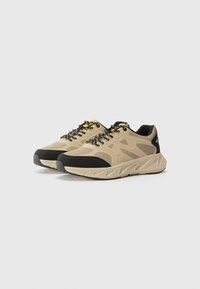 Baskets de sport beiges avec un upper texturé, des accents noirs, une semelle épaisse et des lacets réfléchissants avec une touche de jaune.