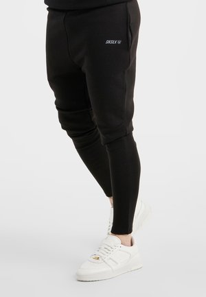 Czarne zwężane spodnie dresowe z logo „SikSilk” na udzie, noszone z białymi sneakersami na neutralnym tle.