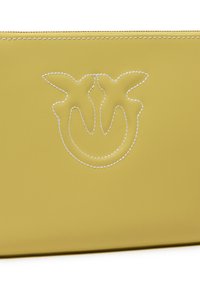 Žuta kožna torbica s utisnutim logotipom, bijelim šavovima, glatkom teksturom i zip zatvaranjem na vrhu. Minimalistički dizajn i kompaktan oblik.