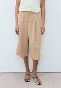 Beige culottes lebar dengan lipitan, terbuat dari kain ringan. Dipadukan dengan sandal minimalis hitam dan atasan putih sederhana.