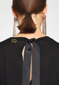 Femme avec une queue de cheval brune portant un haut noir noué avec un ruban à l'arrière et de longues boucles d'oreilles en chaîne argentée, vue de dos.