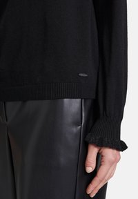 Schwarzer gestrickter Pullover mit geripptem Saum und strukturierten Rüsche-Ärmeln, getragen über schwarzen Lederhosen. Kleines Logo-Detail am unteren Rand.