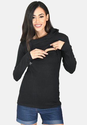 Smiling woman in black long-sleeve top lifts front panel to reveal hidden nursing access, wearing blue rolled-up shorts.
