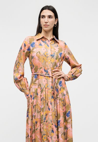 Hosszú sötét hajú nő, aki gombos, hosszú ujjú, virágmintás ruhát visel rózsaszín, kék és sárga árnyalatokban, egyik kezét a csípőjére téve pózol.