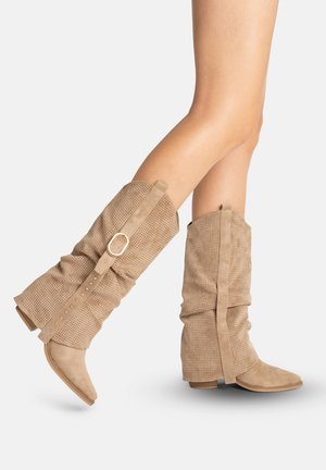 Beine in beigefarbenen Wildleder-Stiefeln mit weitem Schaft bis zur Mitte der Wade, perforiertem Design und dekorativem Riemen mit goldener Schnalle vor einfachem Hintergrund.