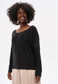 Schwarzes Langarmshirt aus einem weichen Material, mit weitem Ausschnitt und zarten Paillettenakzenten auf der Vorderseite. Kombiniert mit hellen Hosen.