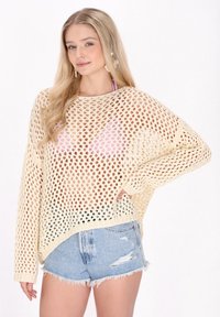 Pulover tricotat de culoare crem, cu un design deschis, cu plasă laxă, având mâneci lungi și decolteu rotund, purtat peste pantaloni scurți din denim.