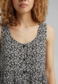 Femme aux cheveux blonds bouclés portant une robe noire sans manches avec un petit motif floral blanc et des boutons noirs à l'avant.