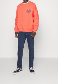 Felpa corallo brillante con vestibilità ampia e polsini a coste, dotata di testo grafico nero. Abbinata a jeans slim blu scuro e sneakers nere.