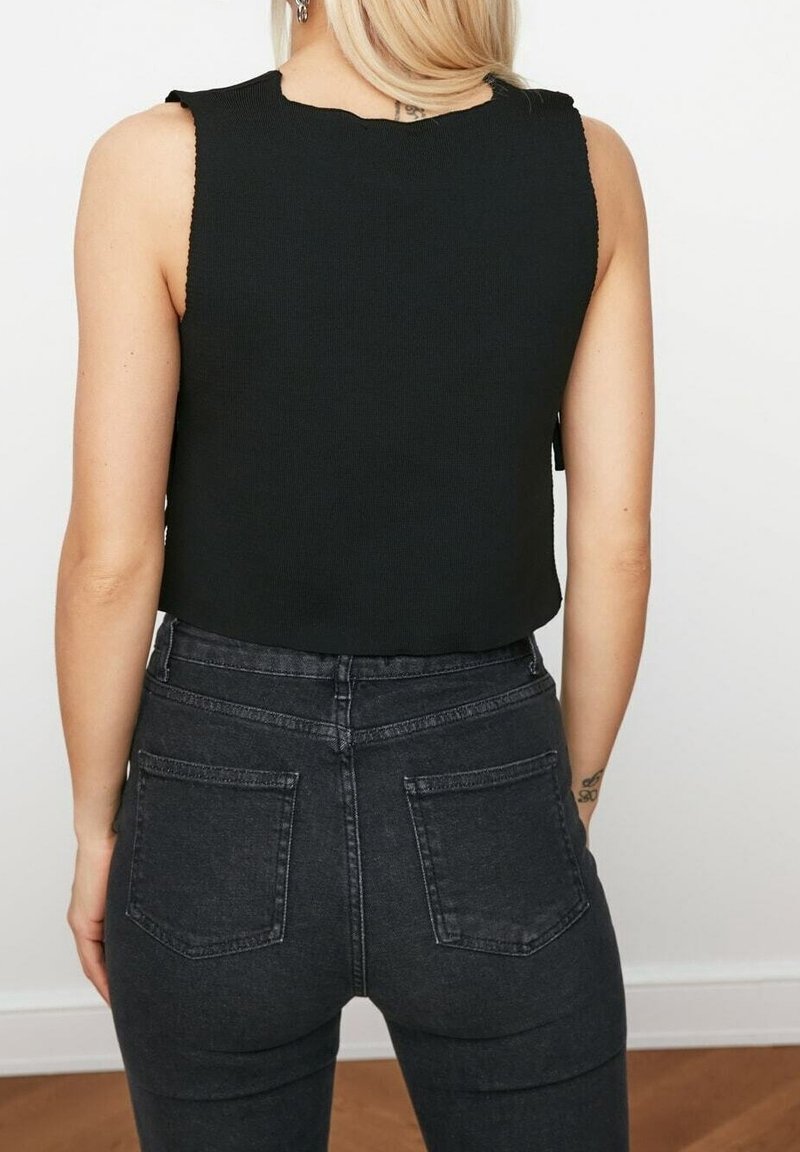 Femme blonde portant un crop top noir sans manches et un jean gris foncé taille haute, vue de dos devant un mur uni.